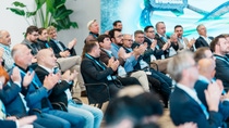 Audience at a symposium conference claps enthusiastically. Attendees, mostly men in business attire, sit in rows with a backdrop of an event banner.