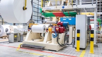 The picture shows a large industrial paper manufacturing or processing machine inside a factory. Several big rolls of white paper are visible, with one roll being lifted by a yellow overhead crane. The machine features various mechanical parts, control panels, and safety barriers. The factory is well-lit with large windows and overhead lights, and the floor has marked safety lines. Nearby, there is a chair and a control desk, indicating an operator’s station. The picture shows a large industrial paper manufacturing or processing machine inside a factory. Several big rolls of white paper are visible, with one roll being lifted by a yellow overhead crane. The machine features various mechanical parts, control panels, and safety barriers. The factory is well-lit with large windows and overhead lights, and the floor has marked safety lines. Nearby, there is a chair and a control desk, indicating an operator’s station.