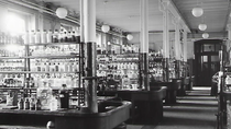 Black and white photo of a large, industrial laboratory with rows of bottles on shelves. The space is lined with sturdy benches.