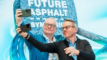 Two men smiling as they take a selfie, standing in front of a sign that reads "Future Asphalt Symposium".