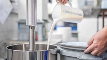 A person pours a thick white liquid from a measuring jug into a large, stainless steel mixer. A person pours a thick white liquid from a measuring jug into a large, stainless steel mixer.