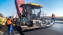 Workers in orange vests operate a paving machine on a sunny day, spreading new asphalt on a road, with a dump truck unloading material. Workers in orange vests operate a paving machine on a sunny day, spreading new asphalt on a road, with a dump truck unloading material.
