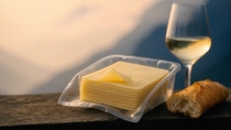 Cheese in plastic packaging tray on a wooden table with white bread and a glass of wine, set against a scenic mountain view in soft evening light.