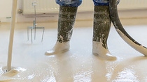 Worker wearing muddy boots is pouring smooth, wet concrete onto a white floor in a bright room, creating a reflective surface. Worker wearing muddy boots is pouring smooth, wet concrete onto a white floor in a bright room, creating a reflective surface.