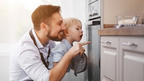 Father and young child in kitchen, peering into oven with curiosity. They are smiling, wearing aprons.