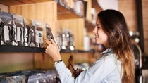 A woman with long hair smiles as she examines a coffee bag on a wooden shelf in a cozy store. She holds glasses in one hand. A woman with long hair smiles as she examines a coffee bag on a wooden shelf in a cozy store. She holds glasses in one hand.