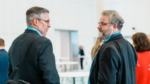 Two men in suits conversing at an indoor conference, smiling and engaged. Conference setting with attendees in the blurred background.
