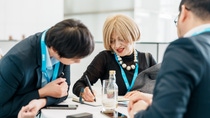 Three people in business attire converse at a round table, smiling and a woman in writing some notes. Name tags suggest a conference setting