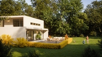 Modern white house with large glass windows, surrounded by lush greenery. A family with a child and dog play on the spacious lawn. Peaceful and sunny setting. Modern white house with large glass windows, surrounded by lush greenery. A family with a child and dog play on the spacious lawn. Peaceful and sunny setting.
