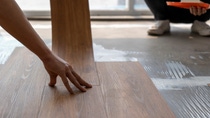 Close-up of hands installing brown wood-like flooring in a sunlit room.