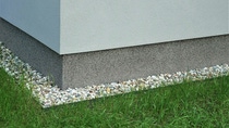 Close-up of a building corner with a smooth, light gray wall. The base is rougher in texture, bordered by small white pebbles and green grass. Close-up of a building corner with a smooth, light gray wall. The base is rougher in texture, bordered by small white pebbles and green grass.