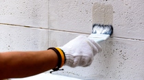 A hand in a white glove paints a textured concrete wall white with a brush. A hand in a white glove paints a textured concrete wall white with a brush.