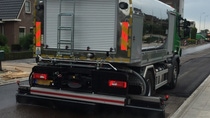 A road construction truck applies a dark liquid coating to a new asphalt road. It is overcast, and nearby houses and a tower are visible in the background. A road construction truck applies a dark liquid coating to a new asphalt road. It is overcast, and nearby houses and a tower are visible in the background.