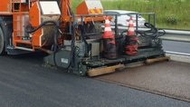 Roadwork scene showing an orange machine laying asphalt on a highway. Traffic cones are stacked, and a worker is visible. A white car passes by.