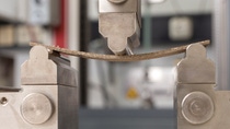 Close-up of a mechanical test where a metal piece is bent between two clamps and a central press. The background is a blurred industrial setting. Close-up of a mechanical test where a metal piece is bent between two clamps and a central press. The background is a blurred industrial setting.