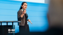 A woman presents confidently on stage with a BASF-branded podium. She wears a black dress and gray cardigan against a blue backdrop.