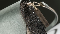 The photo shows a close-up shot of a handbag. The side of the bag is made of coffee beans, brown, the fasteners are golden and the flap is made of green material. On a tag label we can read "seenbrain". The photo shows a close-up shot of a handbag. The side of the bag is made of coffee beans, brown, the fasteners are golden and the flap is made of green material. On a tag label we can read "seenbrain".