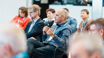 A man in a plaid shirt is holding a microphone, seated among an audience at a conference.