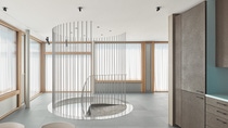 Modern room with sleek, minimalist design featuring a circular, metal spiral staircase, large windows with sheer curtains, and gray cabinets.
Inside the STEP2 unit of the NEST building: View from the kitchen corner to the spiral staircase entry showing a large area of smooth grey flooring. Credit: Zooey Braun