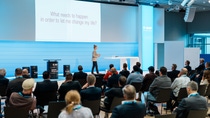 A speaker presents to a seated audience in a modern conference room. The screen reads, "What needs to happen in order to let me change my life?".