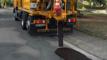 Yellow asphalt repair truck filling potholes on a suburban street. The vehicle dispenses tar, with a distinctive orange cone marking the end of the truck.