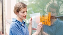 A woman in a blue shirt places an "Open!" sign on a glass window. She looks content and focused. The reflection of the sign is visible on the glass. A woman in a blue shirt places an "Open!" sign on a glass window. She looks content and focused. The reflection of the sign is visible on the glass.