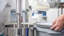 A person pours a thick white liquid from a measuring jug into a large stainless steel mixer. A person pours a thick white liquid from a measuring jug into a large stainless steel mixer.