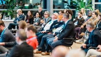 A diverse group of people, seated in a modern conference room, attentively listens to a speaker holding a microphone.
