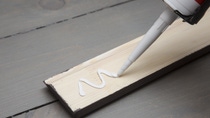 A caulking gun applies a zigzag line of white adhesive onto a light wooden plank on a gray table.