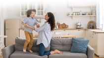 A joyful mother and child play on a gray couch in a cozy kitchen. The child laughs while standing, and sunlight filters through the window. A joyful mother and child play on a gray couch in a cozy kitchen. The child laughs while standing, and sunlight filters through the window.