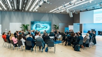 A large group meeting in a modern conference room (D105, Ludwigshafen), with attendees seated in a circle. The room has a futuristic design, including sleek lighting and plants. The atmosphere is collaborative.