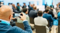 A bald man in a blue sweater photographs a conference with his smartphone. Attendees sit facing a stage, creating a focused and professional atmosphere.