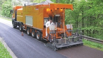 Orange road maintenance truck laying new asphalt on a rural road. A worker adjusts machinery at the back, surrounded by lush green trees.