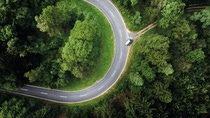 Aerial view of a winding road curving through lush green forested landscape, with a single car traveling the road.