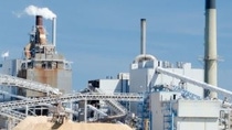 Industrial landscape with a sprawling factory complex under a clear blue sky. Numerous smokestacks and conveyor belts convey a busy, industrial atmosphere.
