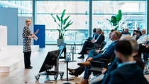 A woman in a patterned dress speaks on stage to an attentive audience in a modern, brightly lit conference room (D105. Ludwigshafen) with large windows and plants.