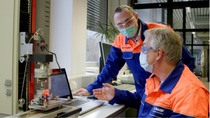 Two masked engineers in BASF uniforms discuss data displayed on a laptop near lab equipment.  Two masked engineers in BASF uniforms discuss data displayed on a laptop near lab equipment.