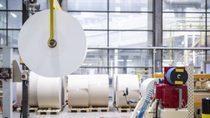 A large paper roll is suspended in a spacious, industrial warehouse with high ceilings and large windows. More rolls are stacked on pallets below. A large paper roll is suspended in a spacious, industrial warehouse with high ceilings and large windows. More rolls are stacked on pallets below.