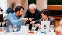 Three men engage in discussion at a dinner table set with wine glasses and a table number card.
