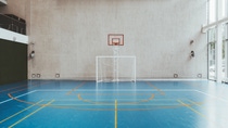 Spacious indoor sports hall with blue flooring, orange and yellow court lines. A soccer goal beneath a basketball hoop against the tall, beige wall. Bright, calm atmosphere. Spacious indoor sports hall with blue flooring, orange and yellow court lines. A soccer goal beneath a basketball hoop against the tall, beige wall. Bright, calm atmosphere.