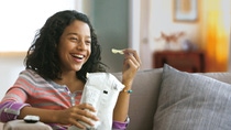 A young girl with curly hair sits on a couch, smiling broadly while holding a potato chip and a snack bag. A young girl with curly hair sits on a couch, smiling broadly while holding a potato chip and a snack bag.