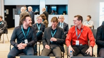 Three men sit in a conference setting, wearing nametags and lanyards. They are smiling and engaged in conversation.
