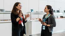 Two women engaged in conversation, holding glasses, wearing lanyards, and standing beside a buffet table in a well-lit conference setting.