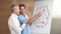 Two men discuss a triangular graph on a flip chart. The chart is labeled with terms like "Stiffness," "Binding Power," and "Robust Resistance." The man on right is wearing a blue polo and the man on the left is wearing a white shirt. Two men discuss a triangular graph on a flip chart. The chart is labeled with terms like "Stiffness," "Binding Power," and "Robust Resistance." The man on right is wearing a blue polo and the man on the left is wearing a white shirt.