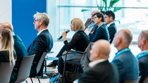 A diverse group of people sitting in a conference room, with a woman in the center holding a microphone, asking a question.