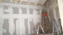 Unfinished room with drywall and visible seams. Two ladders, one with an orange bucket, stand against the walls. Light from a window illuminates the space. Unfinished room with drywall and visible seams. Two ladders, one with an orange bucket, stand against the walls. Light from a window illuminates the space.