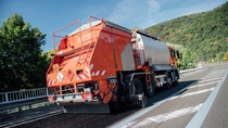 An orange road maintenance truck spreads tar on a highway bordered by metal guardrails and lush green hills, under a clear blue sky. An orange road maintenance truck spreads tar on a highway bordered by metal guardrails and lush green hills, under a clear blue sky.