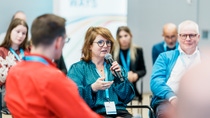 A woman speaks into a microphone in a discussion circle, surrounded by attentive listeners with name tags, creating an engaged and professional atmosphere.