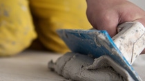 A person kneels on the floor wearing yellow pants, using a blue trowel to spread grey cement. The focus is on the hands and tools. A person kneels on the floor wearing yellow pants, using a blue trowel to spread grey cement. The focus is on the hands and tools.