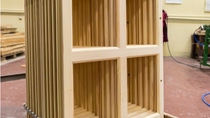 Wooden acoustic diffusers with grid design stand upright in a workshop. They are unpolished and secured with bolts.